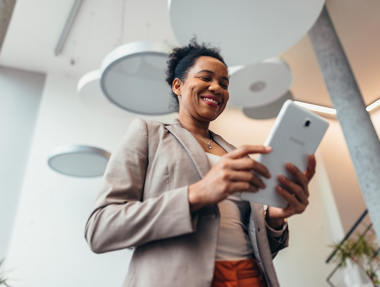 A person smiles and looks at their digital tablet while standing in a modern building lobby.