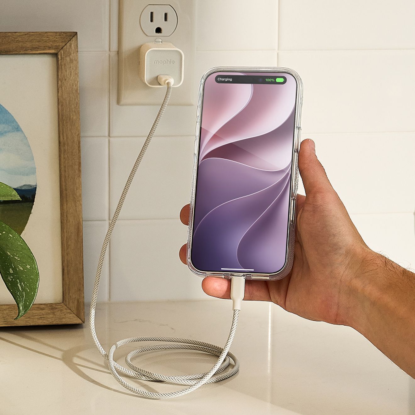 A hand holds a silver iPhone in a clear case while it charges via a white braided USB-C cable connected to a compact white Mophie 35W USB-C wall charger plugged into an outlet. 
