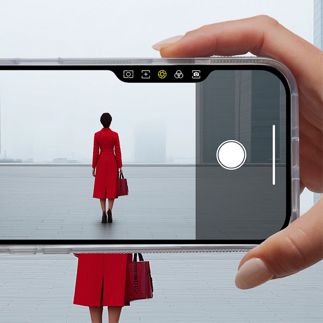 A hand holds an iPhone horizontally in a clear case, using the integrated camera button to frame a photo of a woman in a red coat standing outside a modern building.
