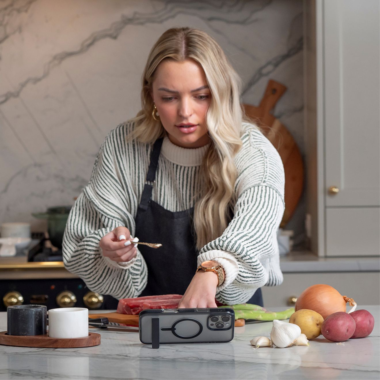 A woman prepares food in a kitchen while a silver iPhone 17 Pro in a clear ZAGG case with a magnetic ring and built-in kickstand is propped horizontally on the counter.