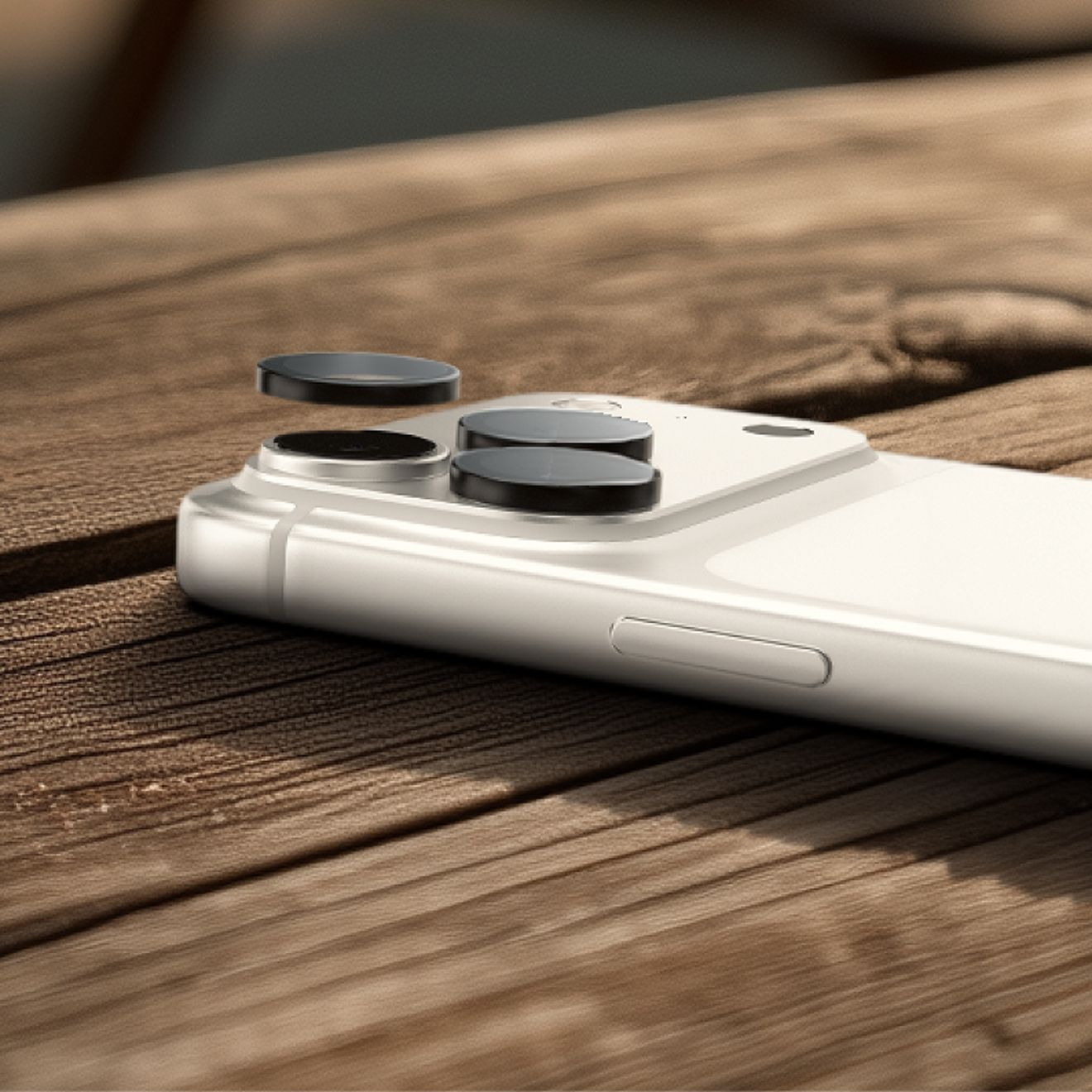Close-up of a silver iPhone Pro on a wooden surface with ZAGG Glass Elite Camera Lens Protectors being lifted from the rear camera lenses using the included lens remover tool.