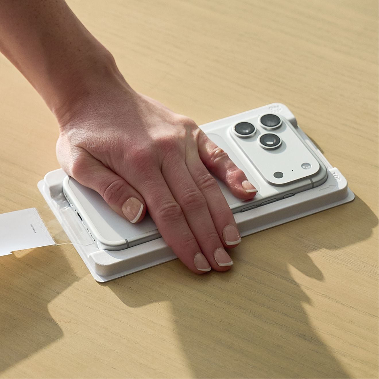 A hand presses down on a silver iPhone positioned in a white PerfectFit™ installation tray on a wooden surface.