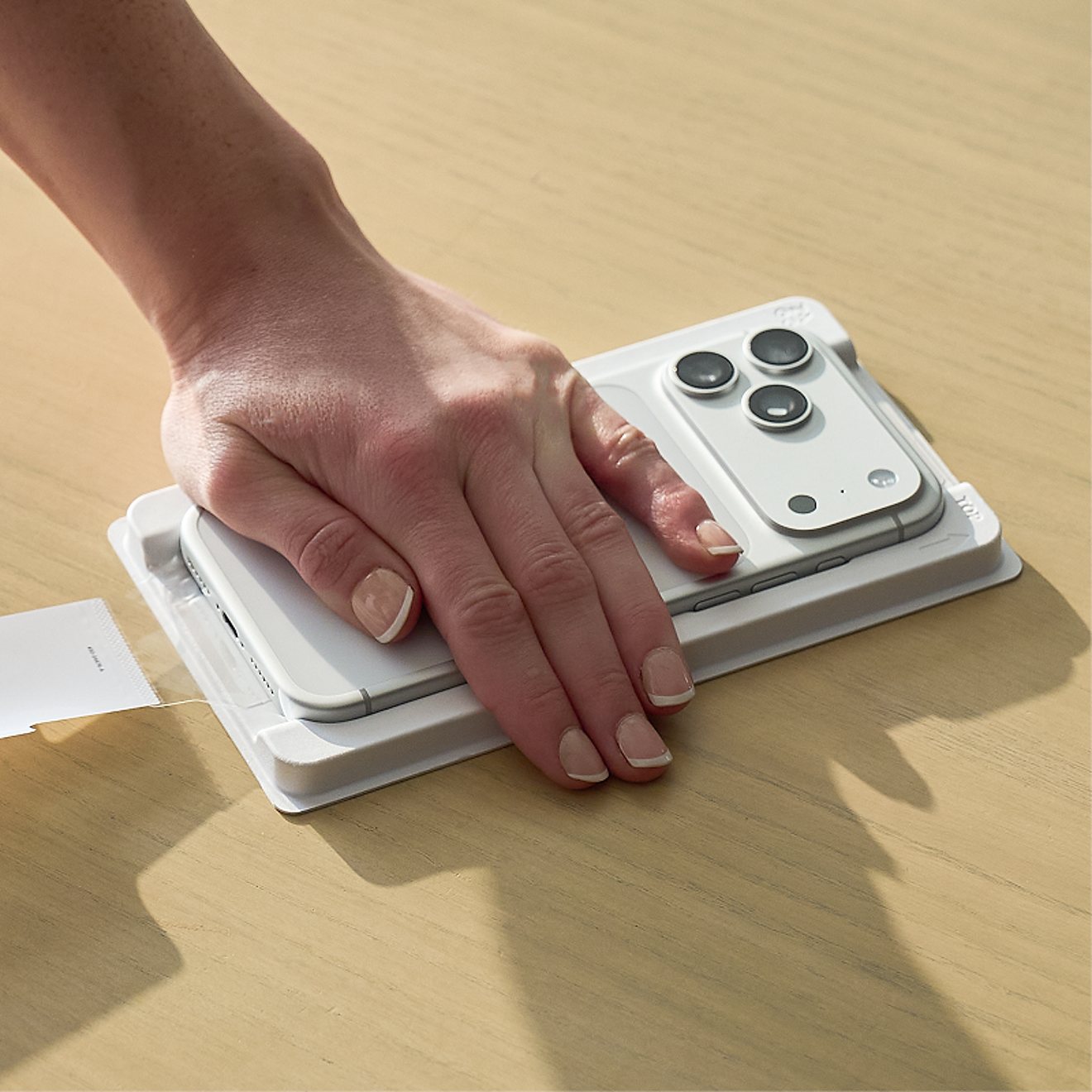 A hand presses down on a silver iPhone positioned in a white PerfectFit™ installation tray on a wooden surface.