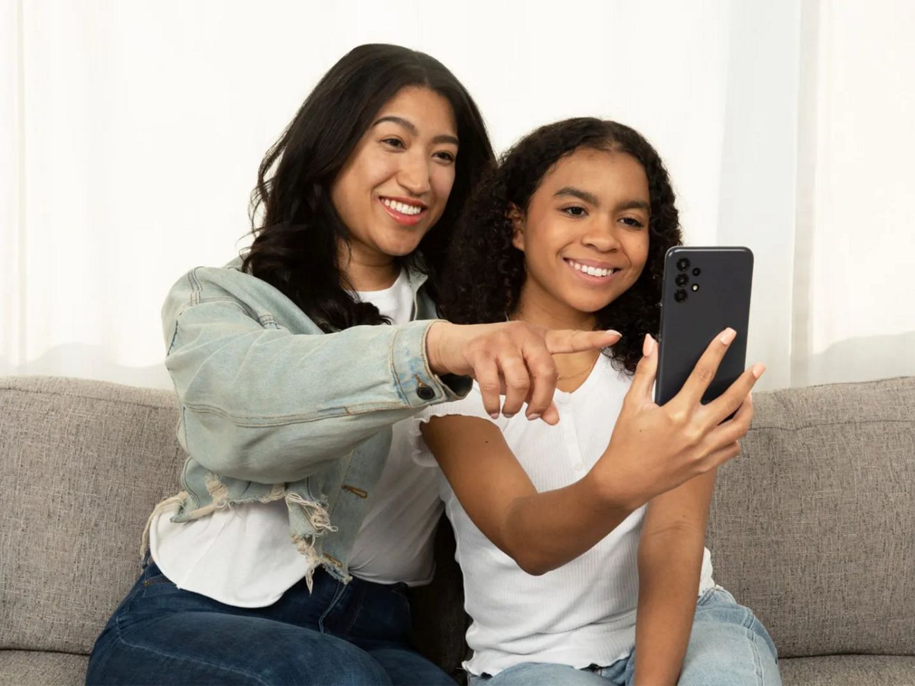 A woman and young girl are smiling as they look at a smartphone. The woman points at the phone display.
