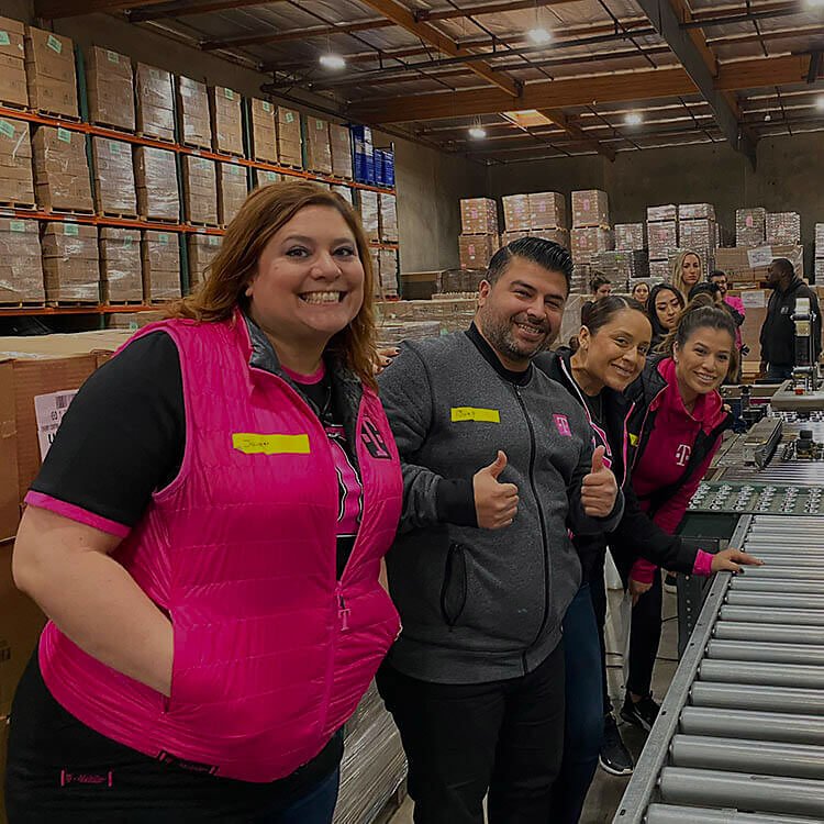 A group of T-Mobile employees smile at the camera while volunteering in a warehouse.