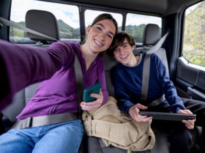 Teenage siblings smile while taking a selfie in the back row of a car.