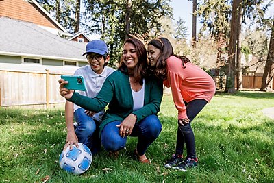 A woman and two kids crouch by a soccer ball in a backyard to take a selfie.