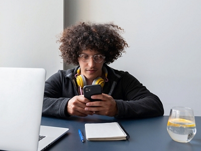 Young man sitting at a desk with headphones around his neck and looking at phone.