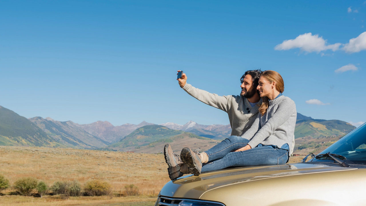 Two people sitting on the hood of a car posing for selfie with desert and mountains in the background
