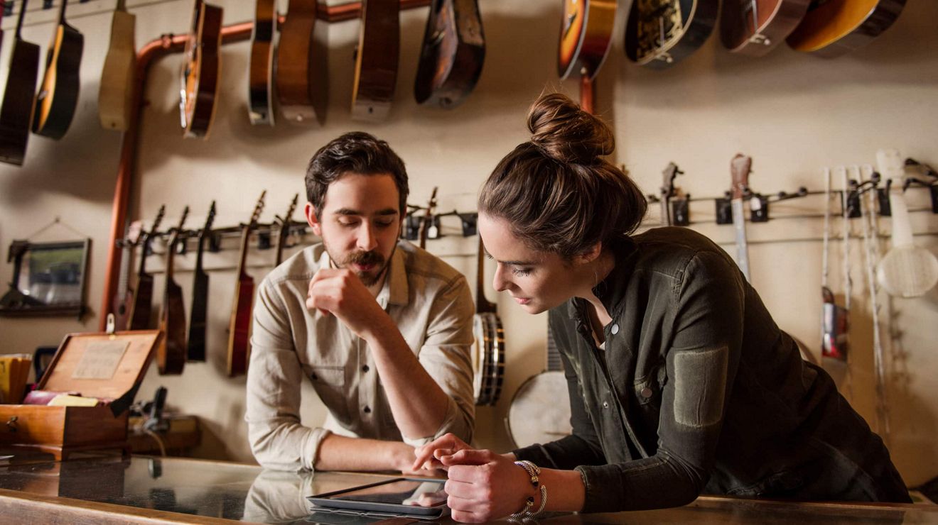 Two employees at a guitar store review inventory on a tablet.