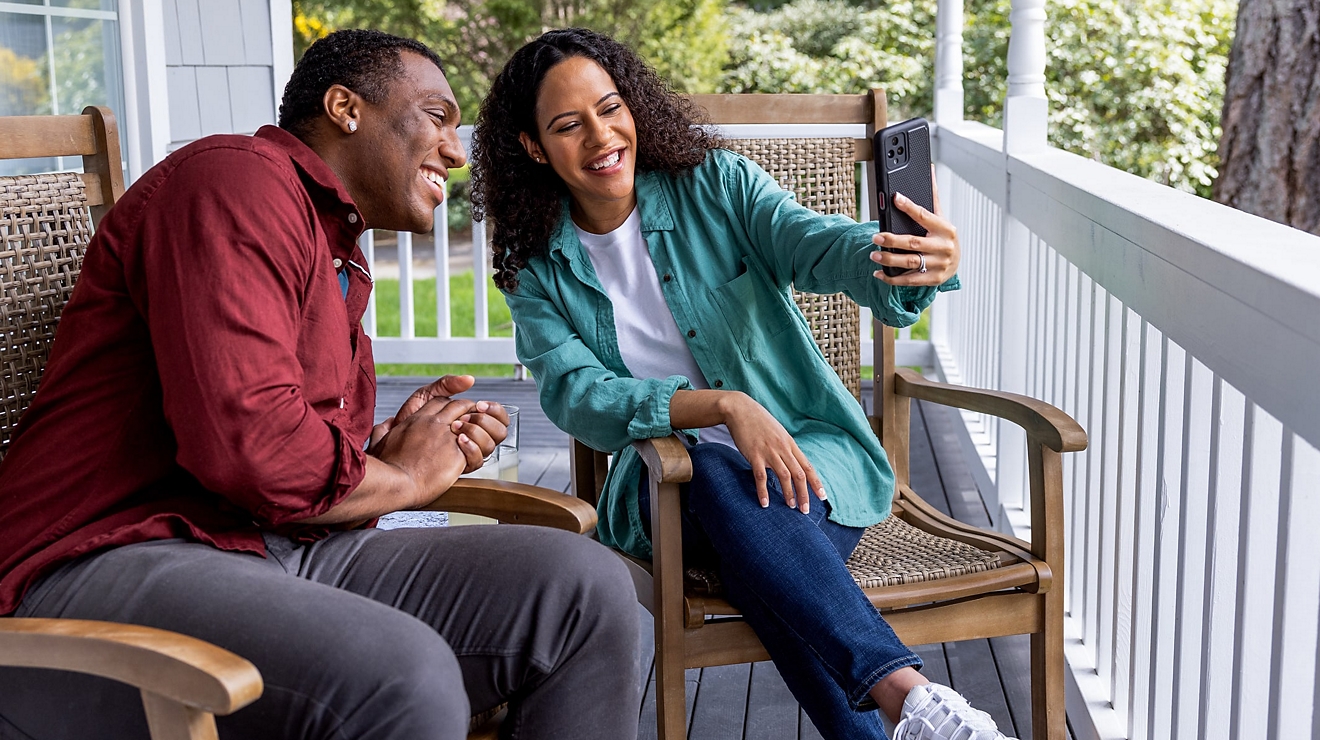 Two people sitting in chairs on a porch while smiling at a phone.