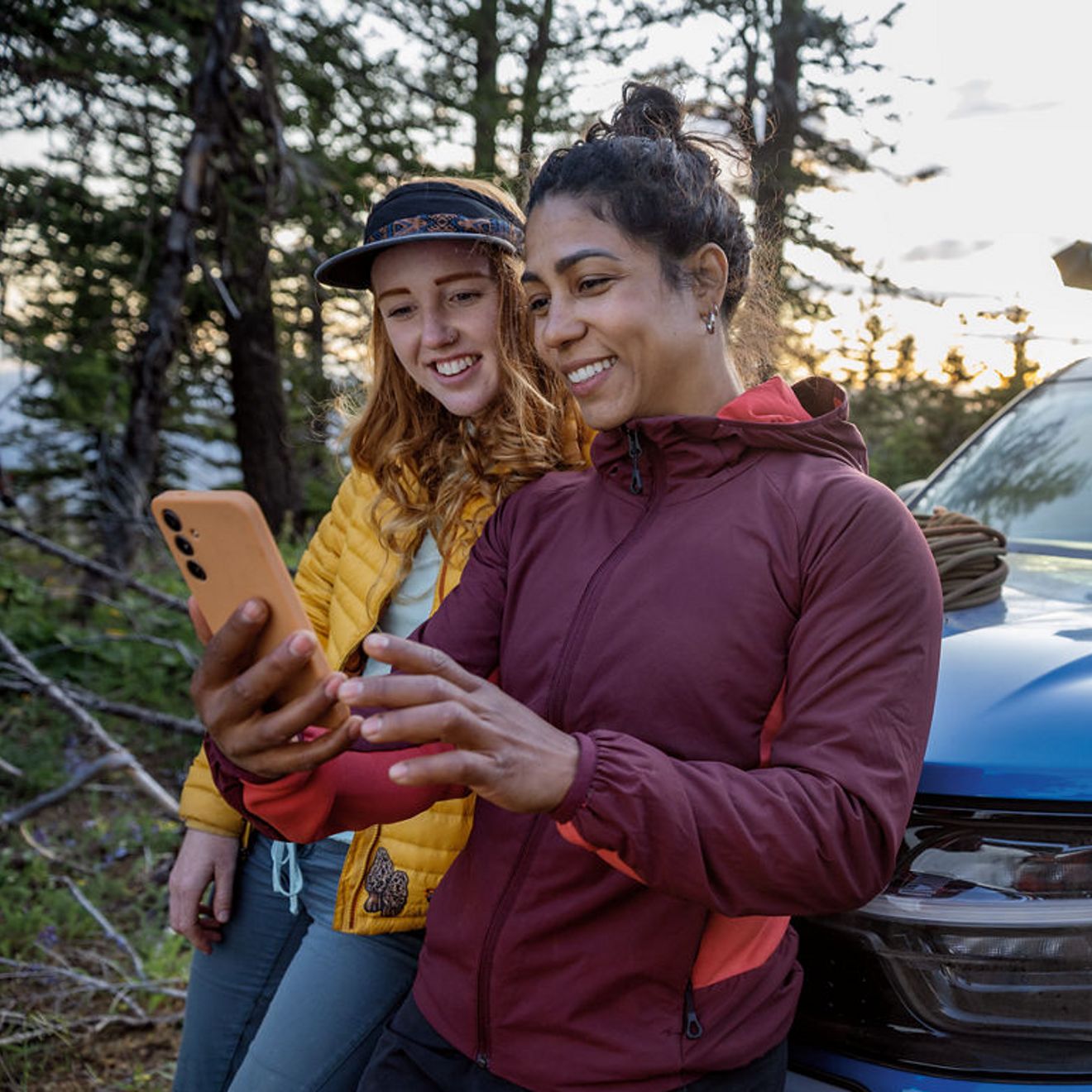 Two women are standing beside an SUV in a forest, smiling as they look at a smartphone together.