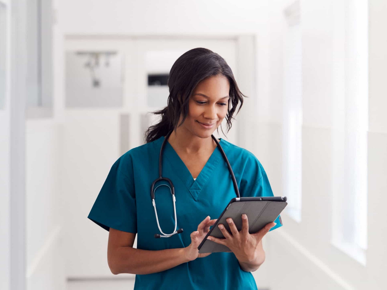 A nurse stands alone in a hospital corridor and checks their digital tablet.