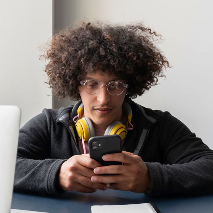 Young man sitting at a desk with headphones around his neck and looking at phone.