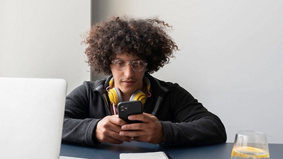 Young man sitting at a desk with headphones around his neck and looking at phone.