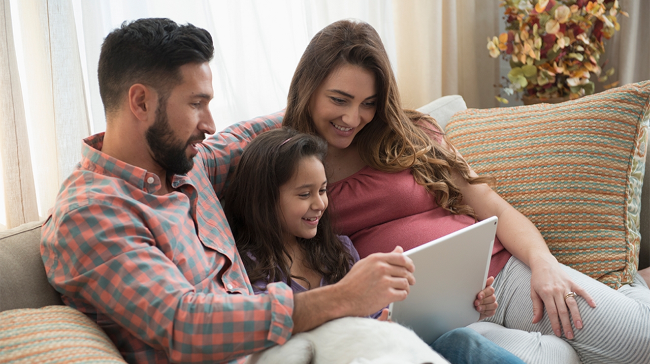 A man, a woman, and a young girl are sitting on a sofa, smiling as they look at a laptop together.