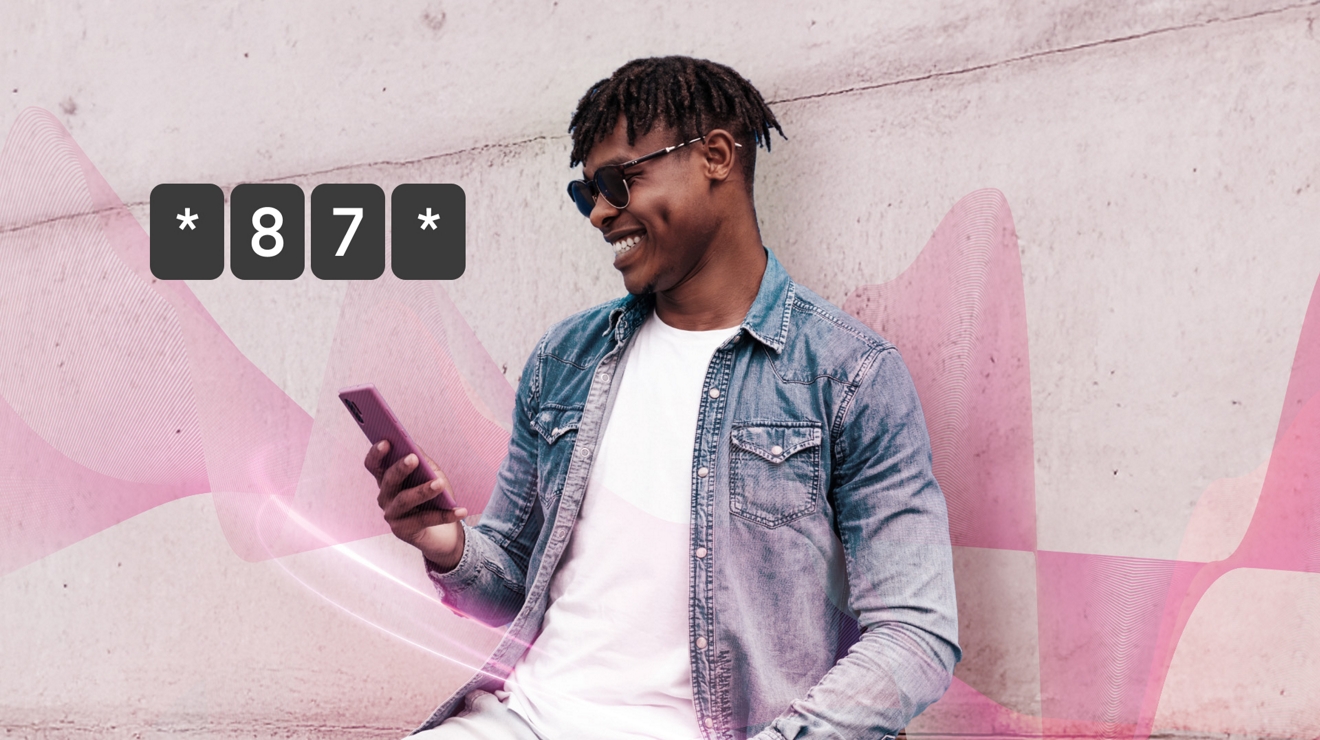 A man in a white t-shirt, denim jacket, and sunglasses smiles at his phone as he uses it. 