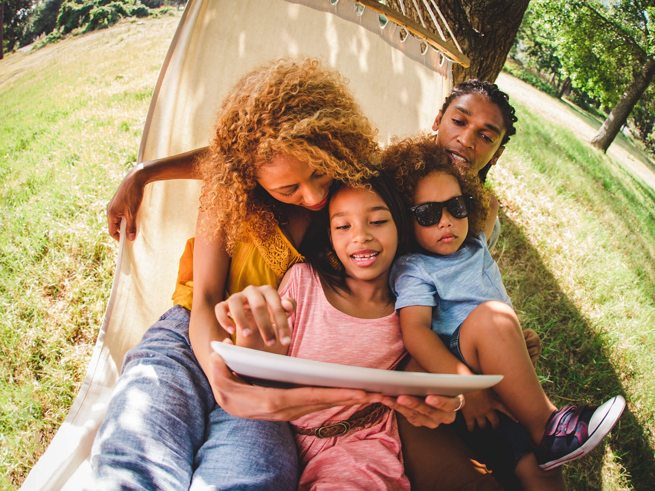 A family of four sits together in a hammock in a grassy park, smiling at a tablet screen.