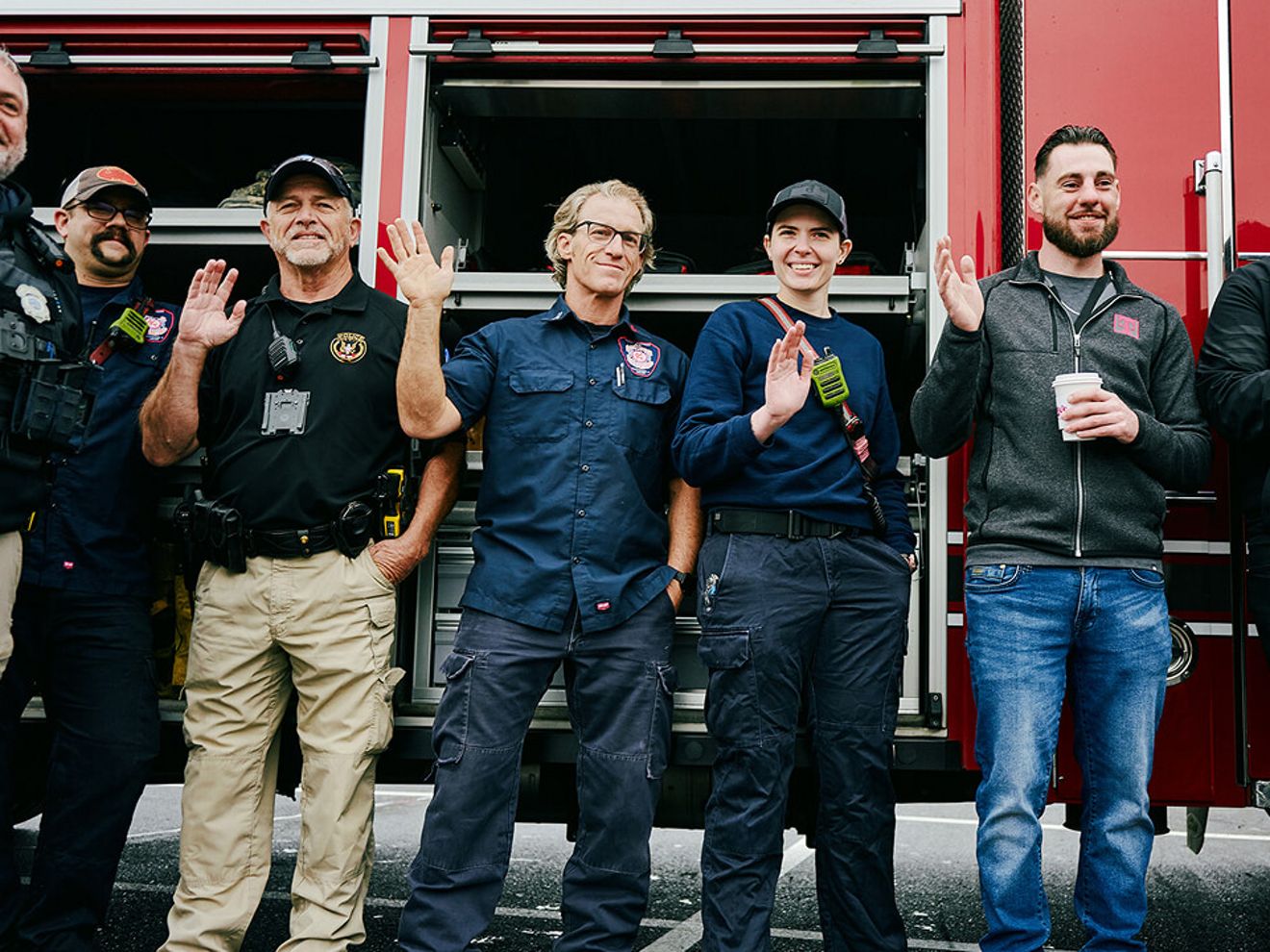 A group of five firefighters and first responders standing in front of a red fire truck, smiling and waving at the camera.