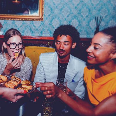 Three friends enjoy snacks and drinks at a lively indoor gathering, seated on a velvet sofa against blue damask wallpaper.