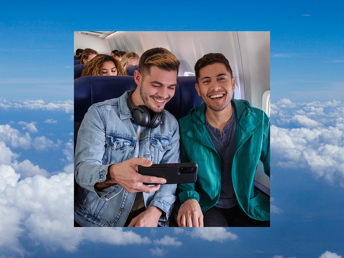 Dos amigos sentados en sus asientos de avión, sonriendo y viendo juntos un video en un smartphone.