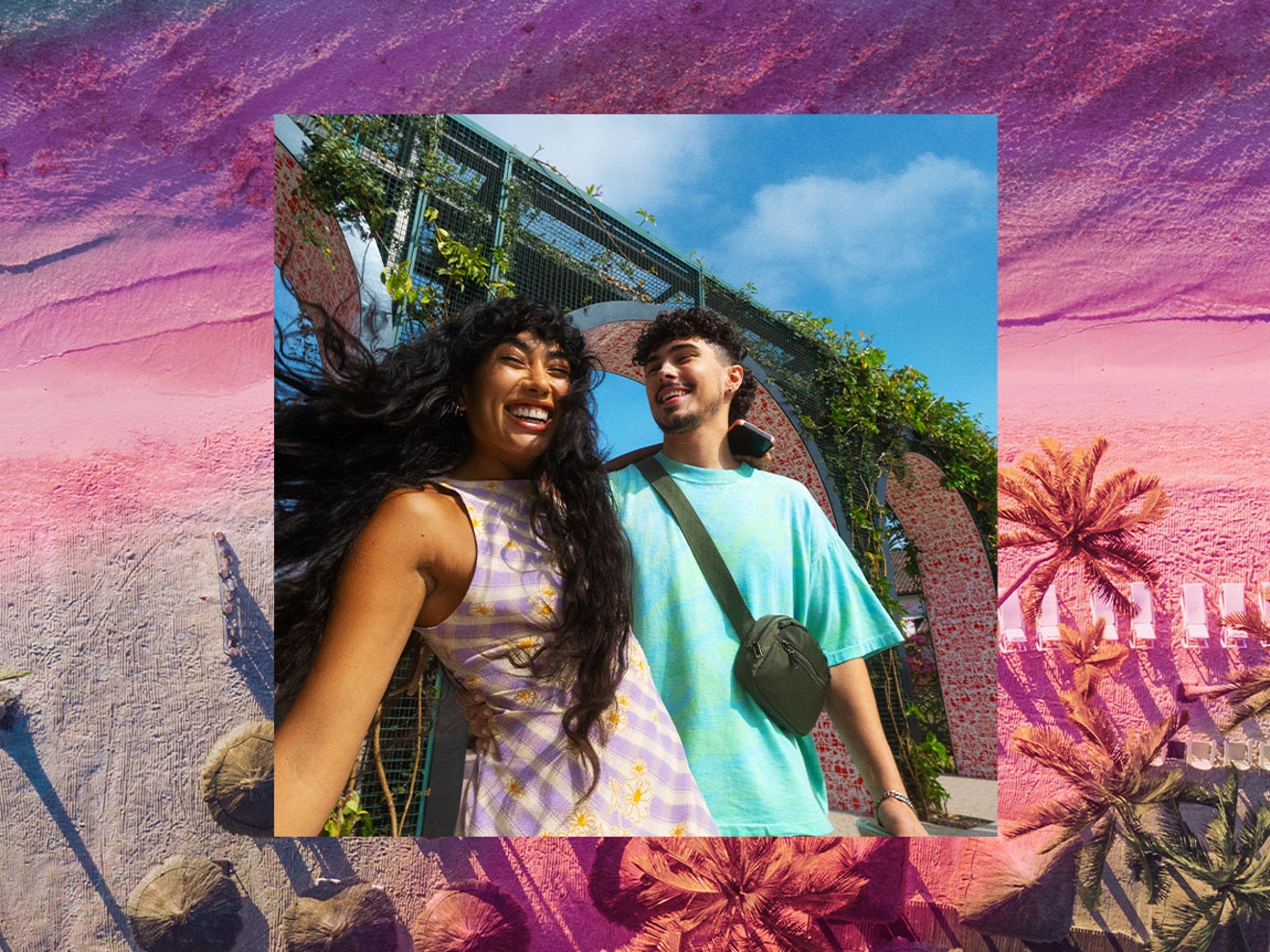 Una mujer y un hombre con trajes coloridos sonriendo al aire libre cerca de un edificio cubierto de vegetación.