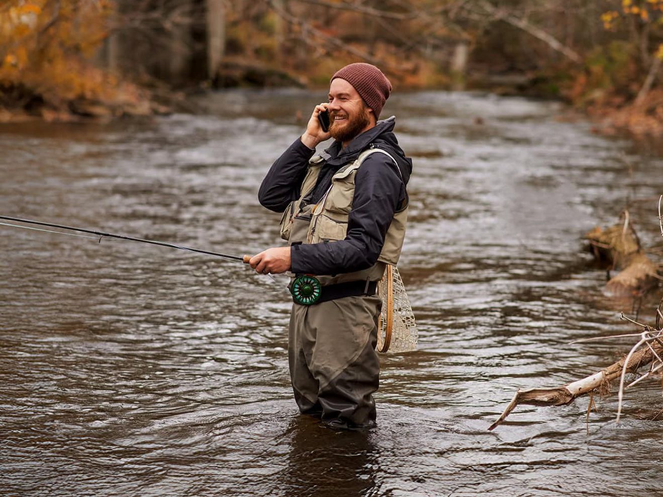 A fly fisherman wearing waders in the middle of a stream holds his fishing pole in one hand while he talks on his phone. 