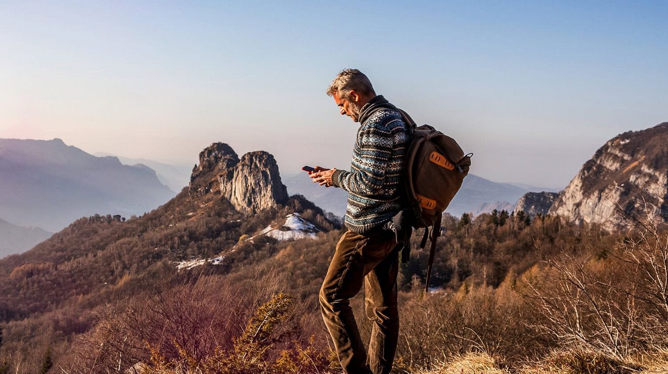 A man wearing a backpack stands in a grassy area high in the mountains, using his phone. 