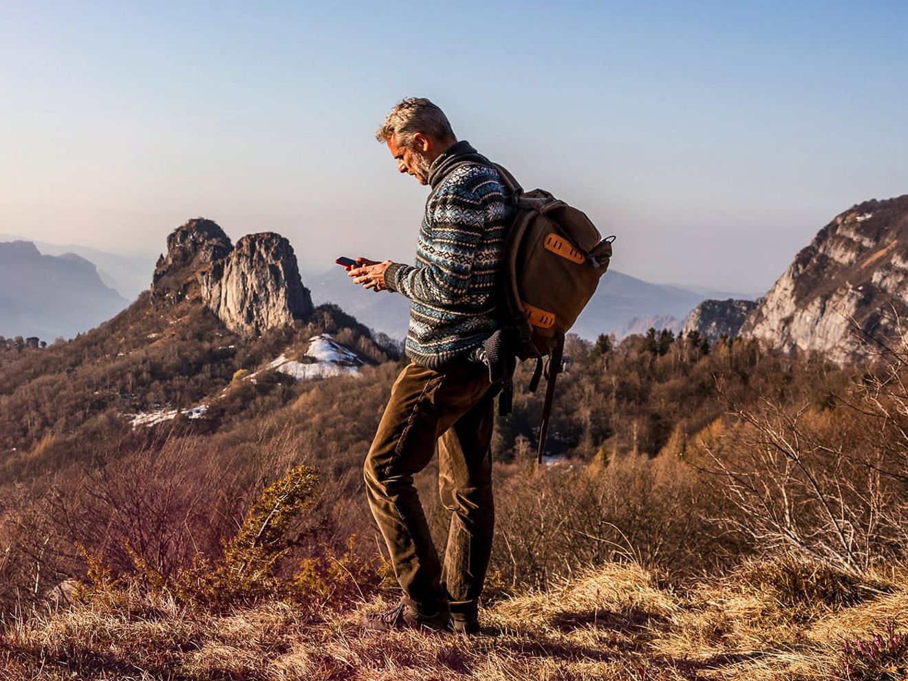 A man wearing a backpack stands in a grassy area high in the mountains, using his phone. 