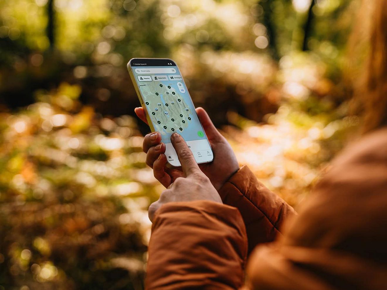 The hands of a person in the woods are shown holding a phone. They are pointing at a pinpointed location shown on a trail mapping app. 