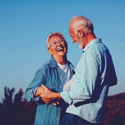 A senior couple laughs together outdoors under a clear blue sky, holding hands and facing each other.