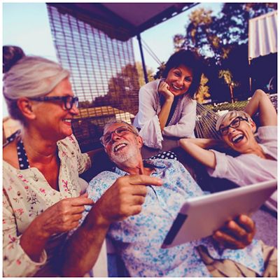 A group of four older adults laugh together on a sunny patio, with one person holding a tablet.