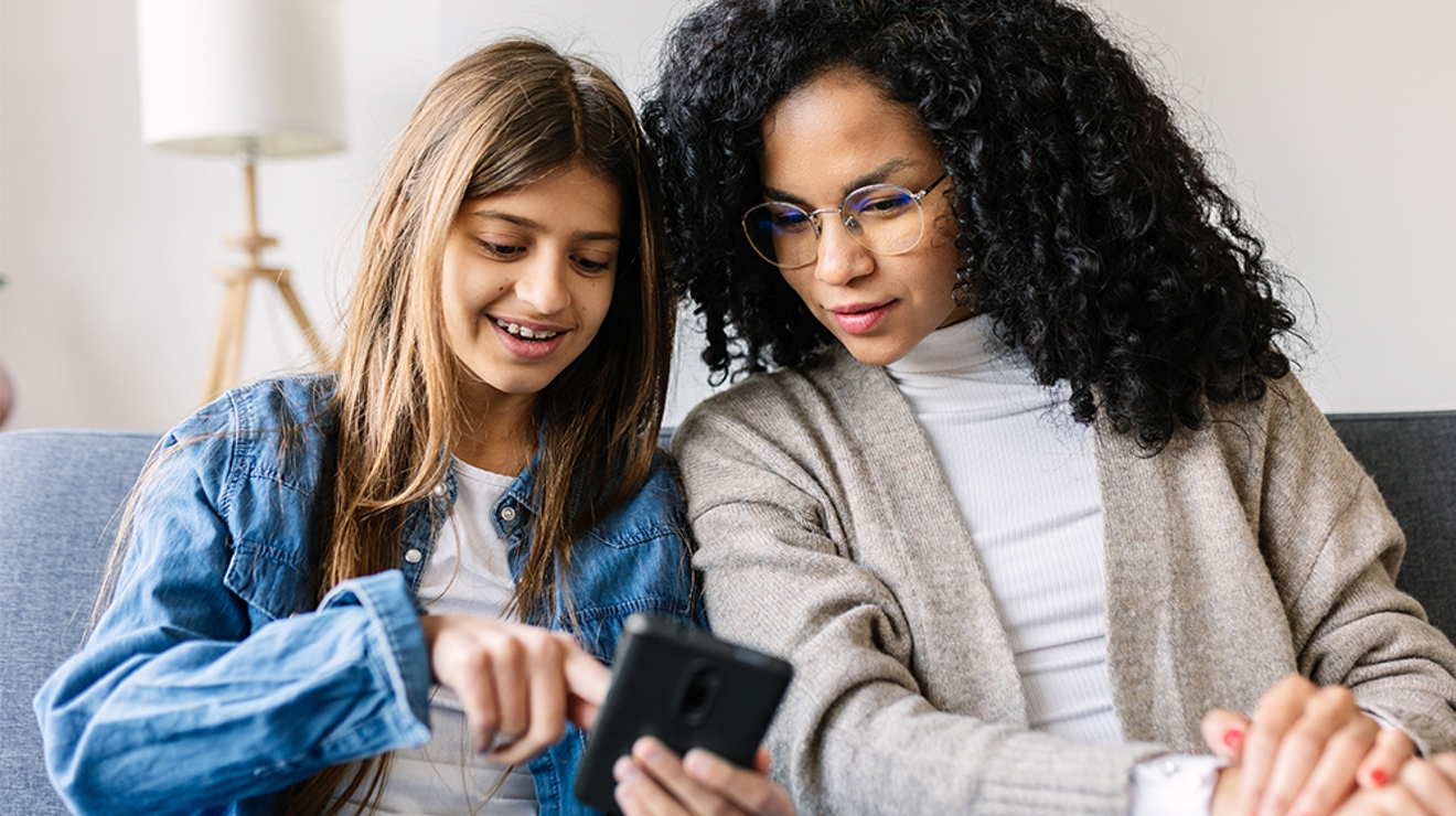 A woman and a young girl are sitting on a sofa. Both are smiling as they look at the girl’s phone.
