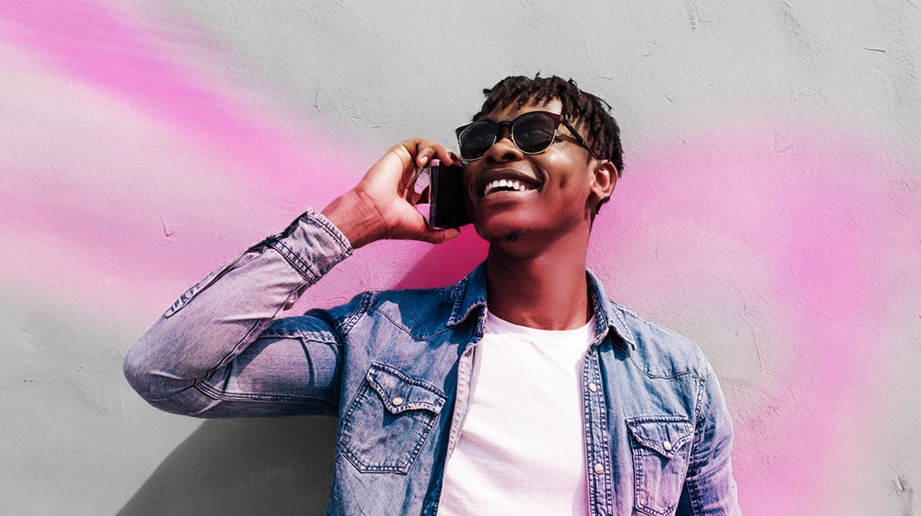A man in a white t-shirt, denim jacket, and sunglasses smiles widely as he holds a phone to his ear and looks up. 