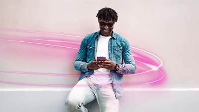 A man in a white t-shirt, denim jacket, jeans, and sunglasses smiles as he looks down at his phone. 