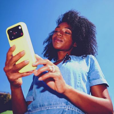 A woman in a denim dress looks down at her phone against a bright blue sky on a sunny day.