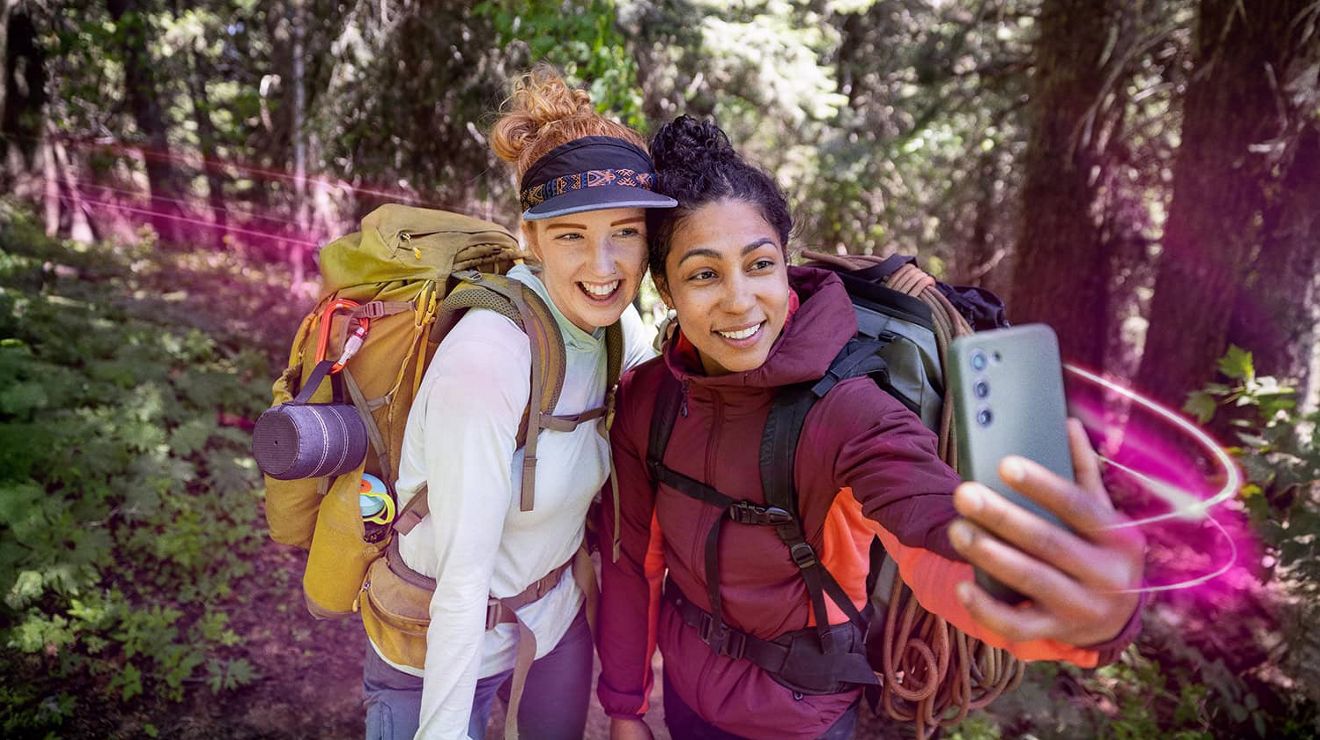 Two women wearing jackets and camping backpacks smile together for a selfie on a wooded trail. 