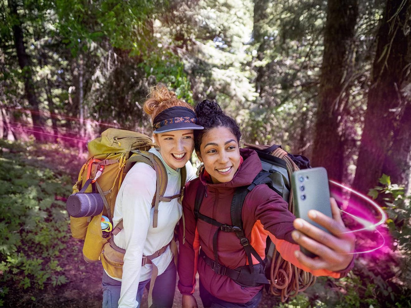 Two women wearing jackets and camping backpacks smile together for a selfie on a wooded trail. 
