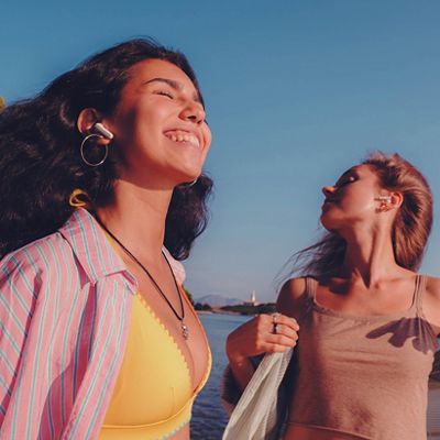 Two women enjoy a breezy day near the water, one wearing wireless earbuds and smiling with eyes closed.