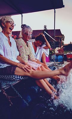 Three women sit on a dock laughing as they splash their feet in the water at sunset.