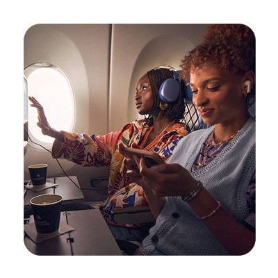 Two women are seated on an airplane. One woman is wearing headphones and looking out the window while the other looks at her phone.
