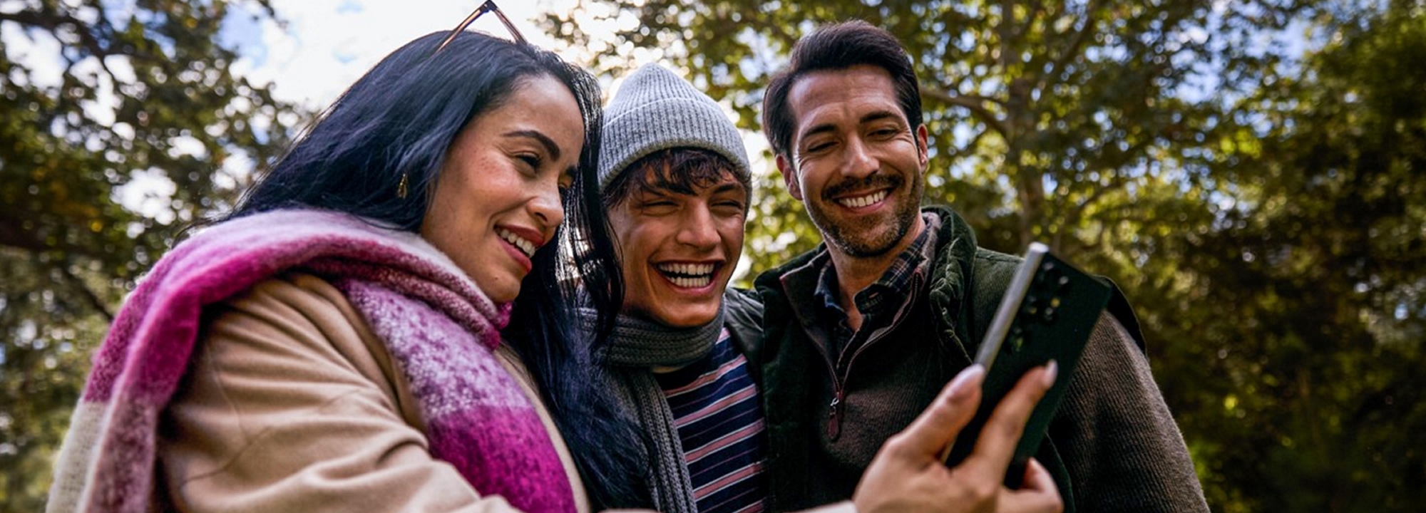 A woman in a magenta and white plaid scarf stands outside, surrounded by a couple of men. All of them smiling at a phone.