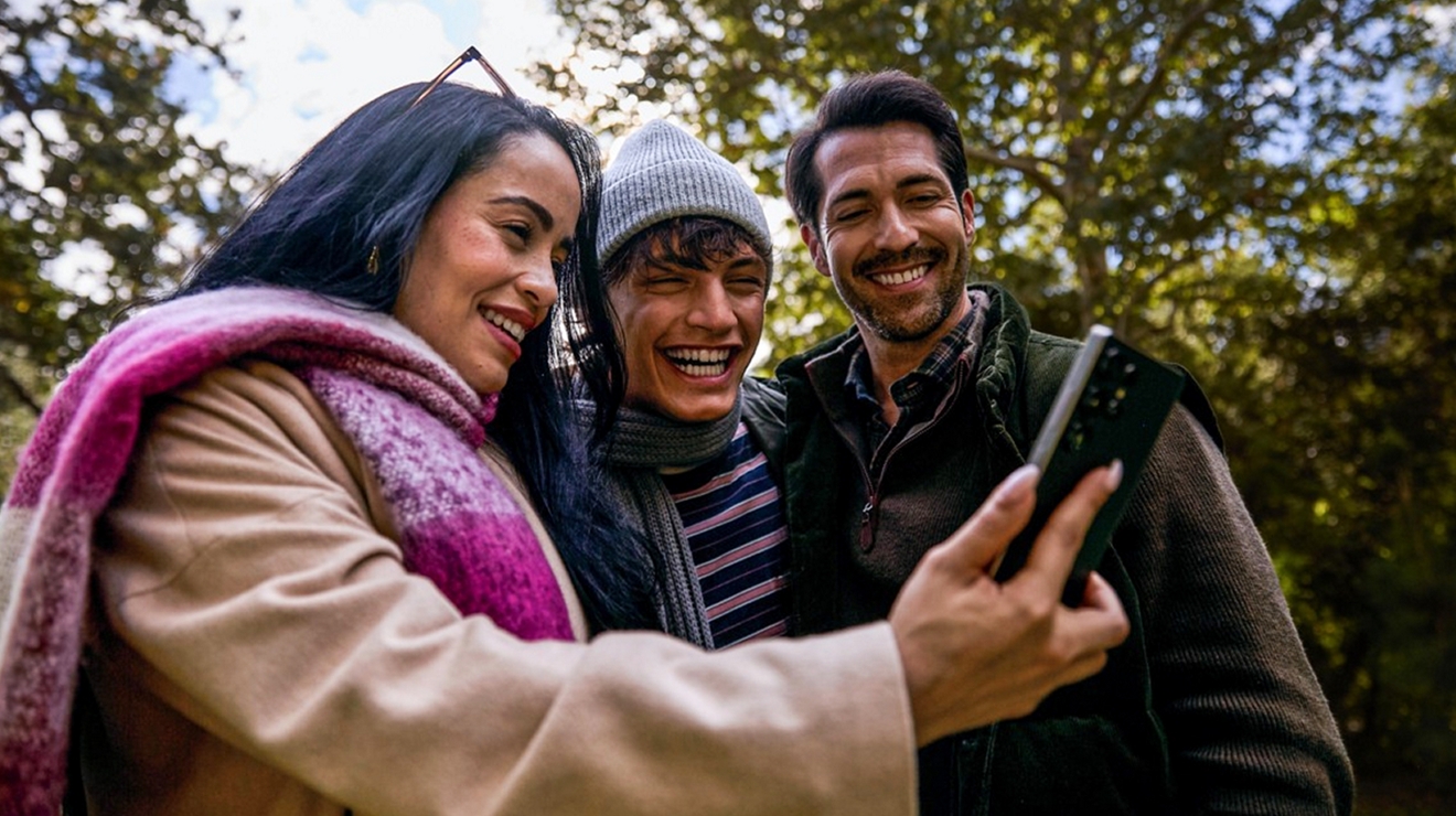 A woman in a magenta and white plaid scarf stands outside, surrounded by a couple of men. All of them smiling at a phone.