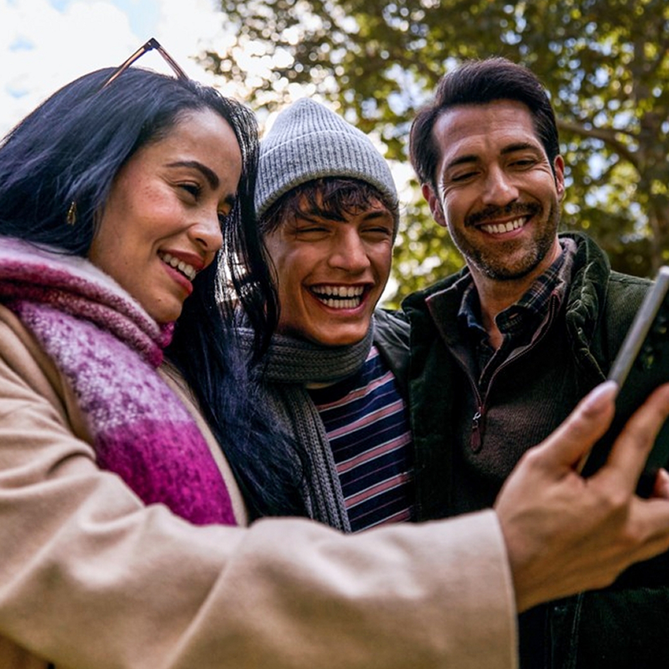 A woman in a magenta and white plaid scarf stands outside, surrounded by a couple of men. All of them smiling at a phone.