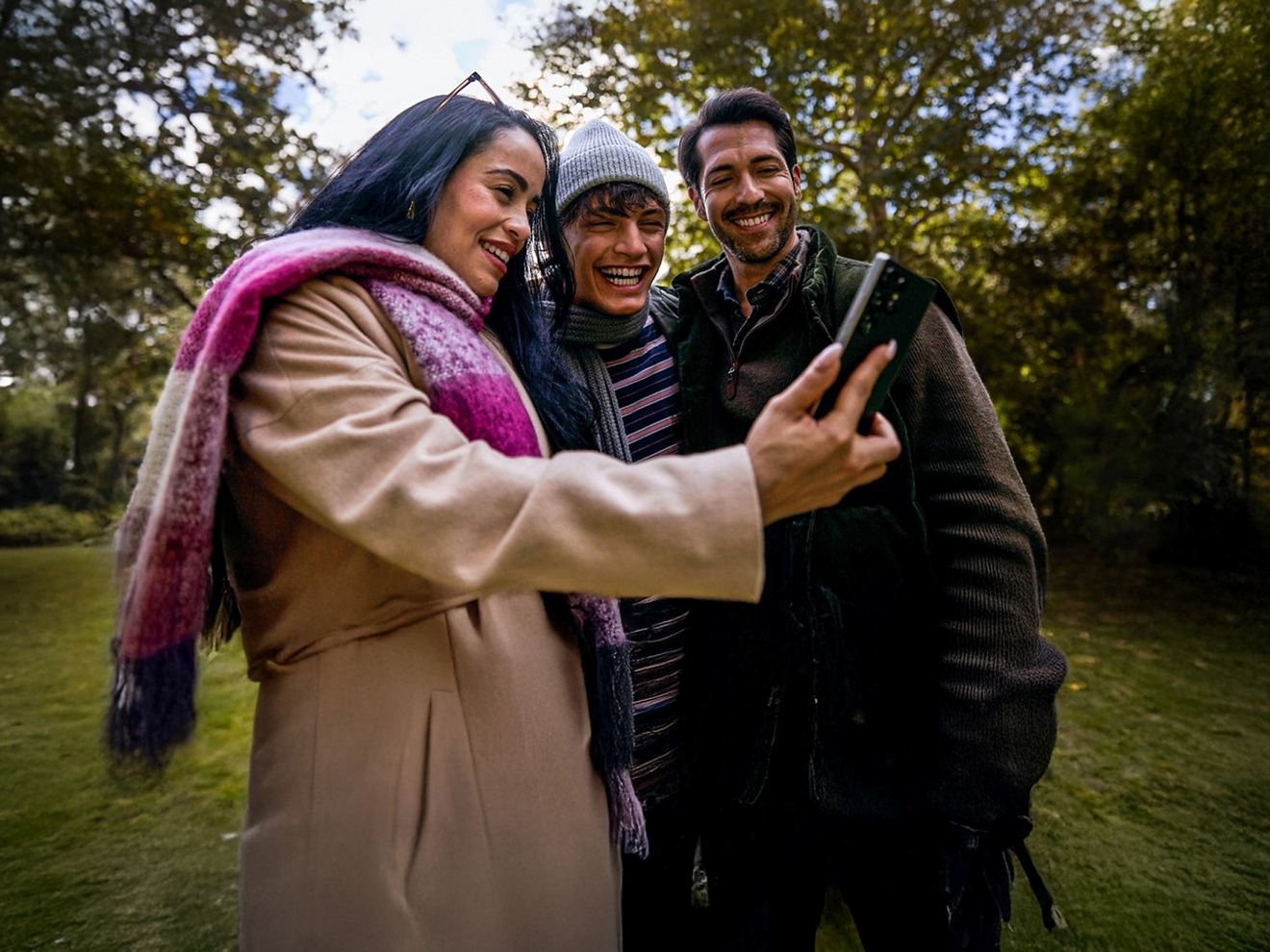 A woman in a magenta and white plaid scarf stands outside, surrounded by a couple of men. All of them smiling at a phone.