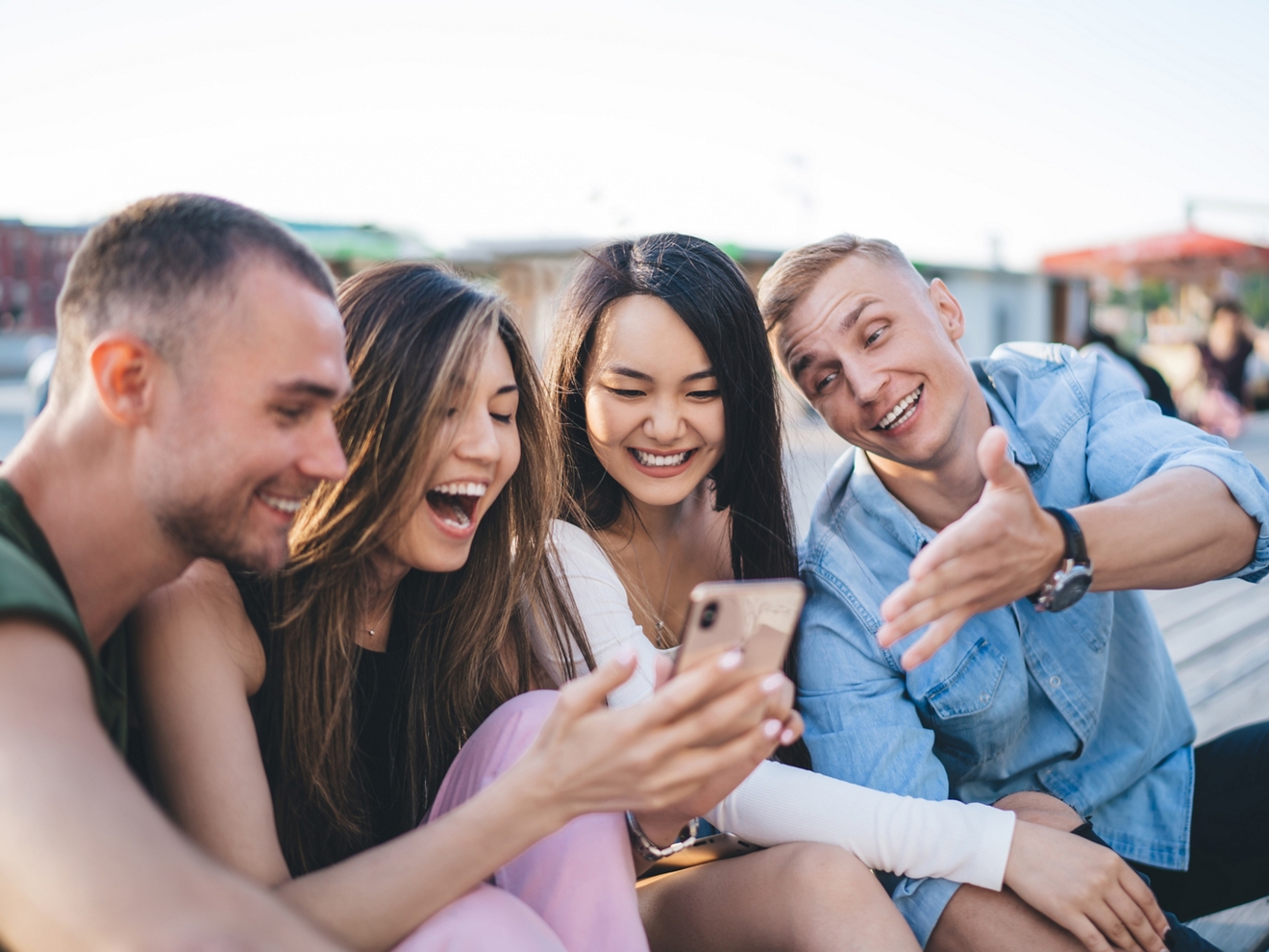 ​​A group of four friends sitting outside laugh and smile while looking at one woman’s phone.​ 