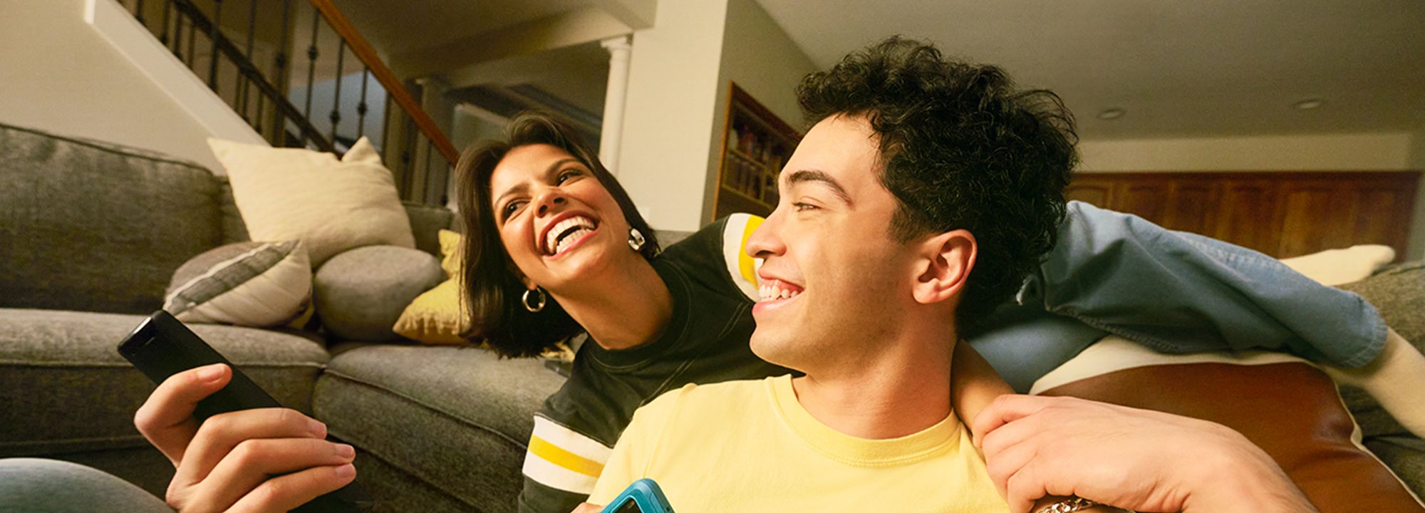 A couple joking around while sitting on a couch, with their phones on their hands.