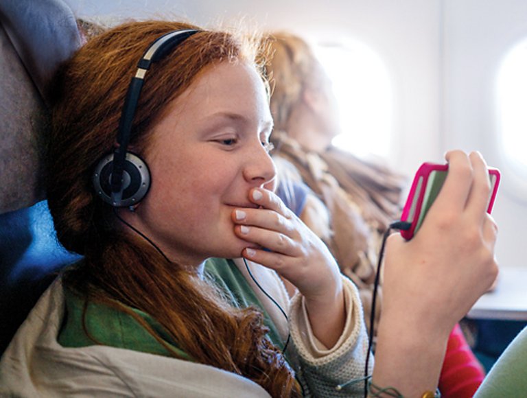 On an airplane, a young girl wearing headphones smiles at her phone. 