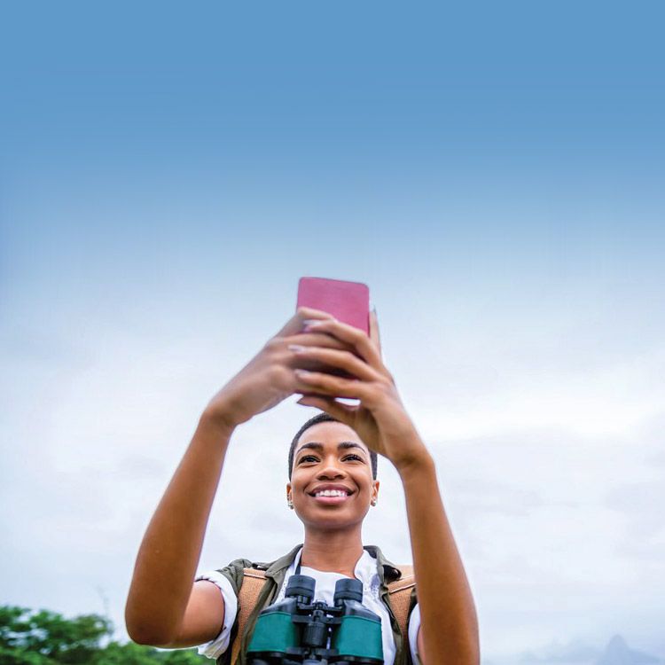 A woman stands outside with a backpack and binoculars around her neck and smiles up at her phone.