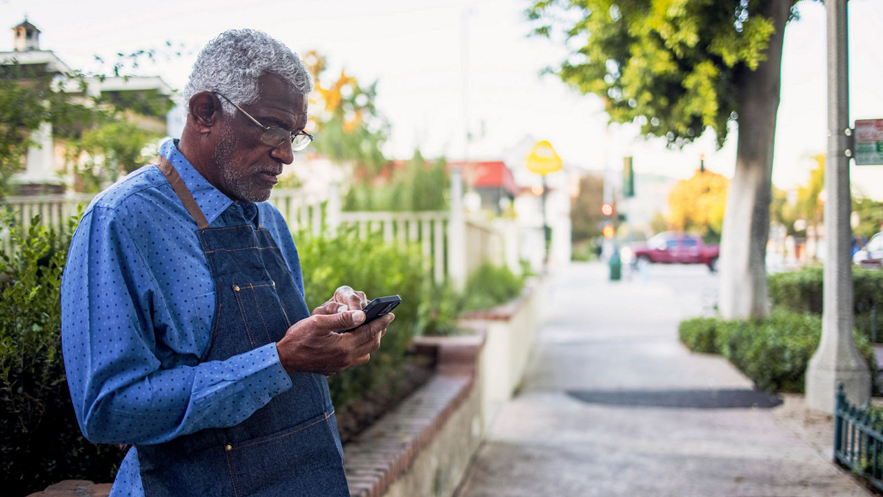 Older man, wearing a craftsman’s apron, texting on smart phone on a neighborhood sidewalk